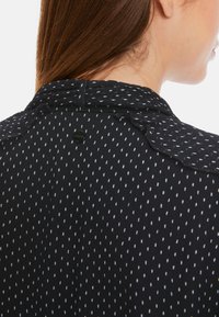 Schwarze Bluse mit weißem Punktmuster, ausgestattet mit einem Kragen mit gewellten Kanten und einem kleinen Knopf auf der Rückseite. Weicher Griff, leichtes Material.