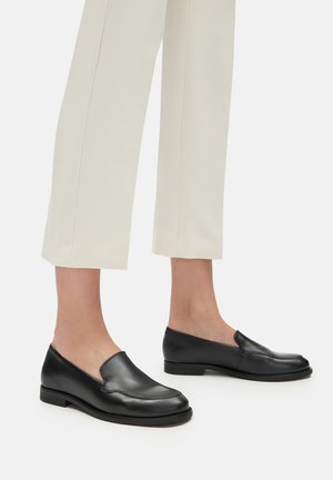 Black leather slip-on loafers worn with beige cropped straight-leg pants on bare feet, shown from side on white background.