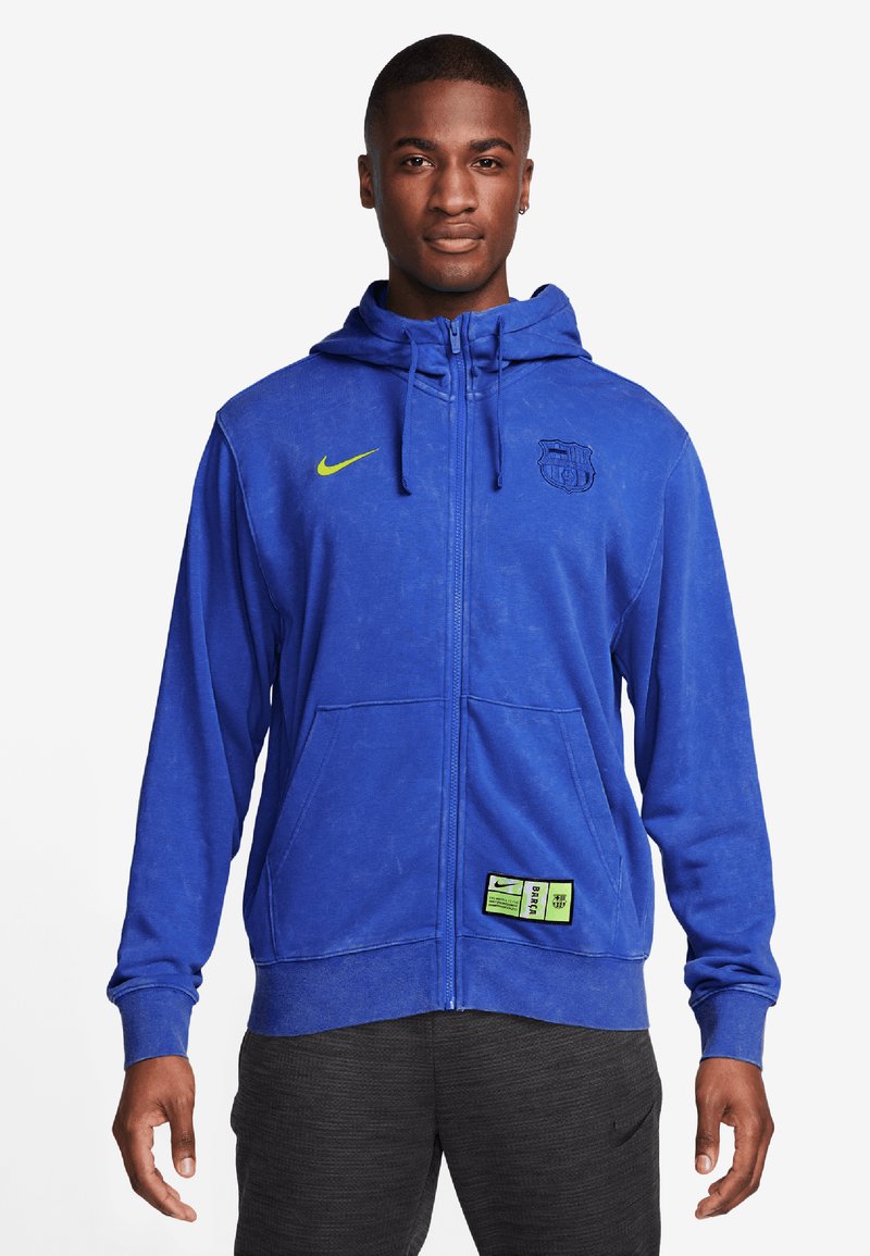 Sudadera azul con cremallera, hecha de tela suave, con un logo de Nike y un emblema del Barcelona, con dos bolsillos y una etiqueta verde en la parte inferior.