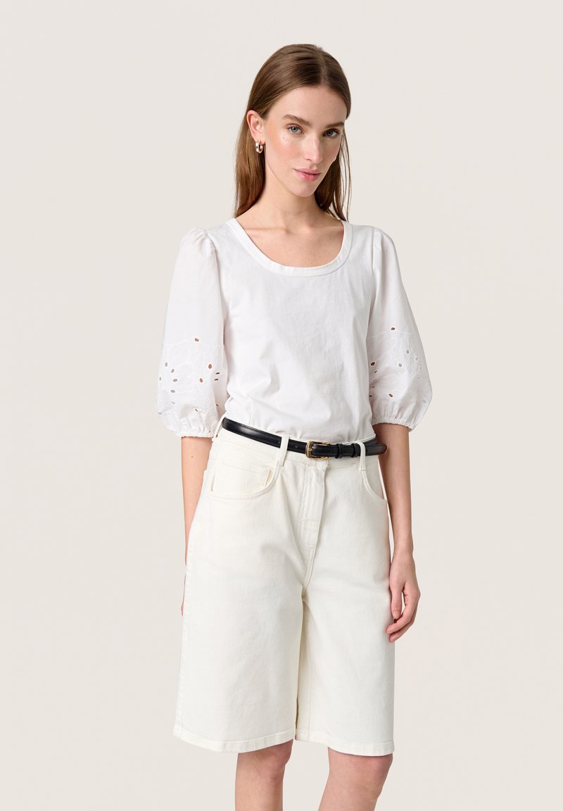 Blouse en coton blanc avec des manches bouffantes et des détails en broderie à œillets. Accordée avec un short bermuda en denim blanc et une ceinture noire.