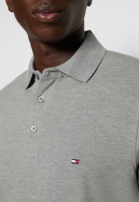 Polo gris en tissu texturé, doté d'un col et de trois boutons, agrémenté d'un petit logo rouge, blanc et bleu sur la poitrine.