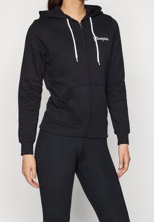 Osoba ubrana w czarną bluzę z kapturem zapinaną na zamek, z białymi sznurkami i logo "Champion" na piersi, oraz czarne legginsy, stojąca na jednolitym tle.