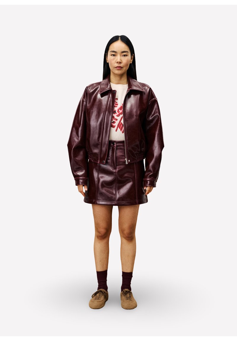 Bordeaux faux leren jack met een ritssluiting, bijpassende rok en lichte trui met een rode grafische design. Bruine schoenen en donkere sokken.