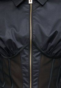 Κοντινό πλάνο ενός μαύρου ενδύματος με χρυσό φερμουάρ στο κέντρο, που διαθέτει δομημένα cups και διάφανα πλαϊνά πάνελ.