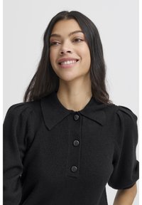 Juodas megztas viršus su klasikiniu apykakle, trumpomis pūstomis rankovėmis ir trimis priekinėmis sagomis. Audinys yra minkštas, su tekstūruota išvaizda.