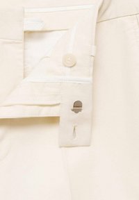 Gros plan d'un pantalon couleur crème avec une taille ouverte montrant un bouton, une fermeture éclair et une fermeture métallique à crochet et barre.