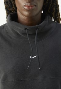 Gros plan d'une personne portant un pull en polaire noir Nike avec un col montant et des cordons, montrant les lèvres et une partie des cheveux tressés.