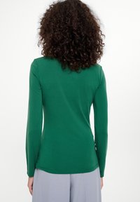 Zielona bluzka z długim rękawem o dopasowanym kroju, gładkiej fakturze, okrągłym dekolcie i bocznymi rozporkami, w połączeniu z jasnoszarymi spodniami.
