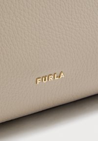 Superficie de cuero beige texturizado con pequeñas letras metálicas doradas que forman la palabra "FURLA", centradas cerca del borde inferior.