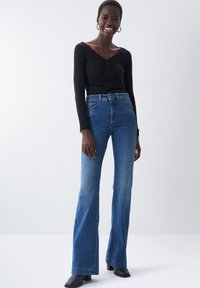 Jeans a palazzo blu in denim ad alta vita con lavaggio medio, design a cinque tasche e chiusura con bottone, abbinati a un top nero a coste a maniche lunghe.