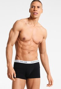 Schwarze Shorts mit einem weißen Bund, der den Text "TOM TAILOR" trägt. Der Stoff wirkt glatt und eng anliegend, wodurch die Konturen des Körpers betont werden.