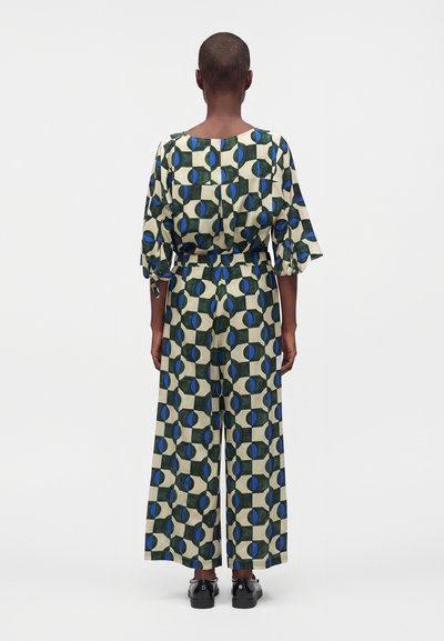 Personne debout de dos, vêtue d'un haut à motifs assortis et d'un pantalon large beige, vert et bleu, avec des chaussures noires.