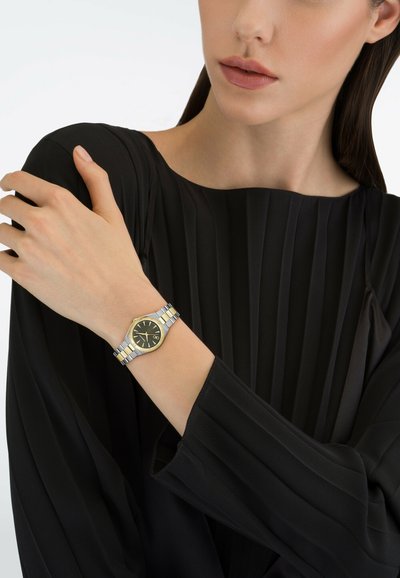 Vrouw draagt een zwart geplooid shirt met lange mouwen en toont een tweekleurig zilveren en gouden horloge aan haar linkerarm.