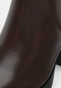 Close-up de um sapato de couro castanho escuro com textura suave e costura subtil perto do tornozelo, apresentando um salto preto baixo.