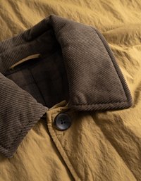 Rjav kordurojski ovratnik na gorčično rumeni zunanji plasti, z enim črnim gumbom in teksturirano tkanino z nagubanim zaključkom.