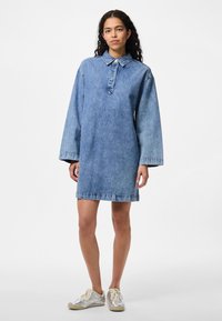 Denimklänning i ljusblått, med krage och knäppning fram, långa ärmar och en rak silhuett. Har en nederkant med rå kant.