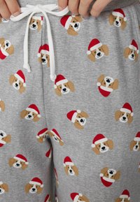 Grijze joggingbroek met een herhalend patroon van cartoonhonden die rode Santa-mutsen dragen, met een witte koord aan de taille.