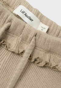 Tessuto beige a costine con un bordo arricciato, che presenta un'etichetta bianca con la scritta "Lil' Atelier" e informazioni sulla taglia. Morbido al tatto, adatto ai bambini.