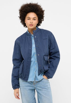 Dunkelblaue gesteppte Jacke mit rundem Kragen und Druckknöpfen. Mit elastischen Bündchen und zwei vorderen Taschen. Über einem Jeanshemd getragen.