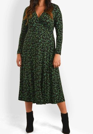 Vrouw draagt een zwart midi-jurk met lange mouwen en een groene bloemenprint, gecombineerd met zwarte enkellaarzen, staand tegen een effen lichte achtergrond.