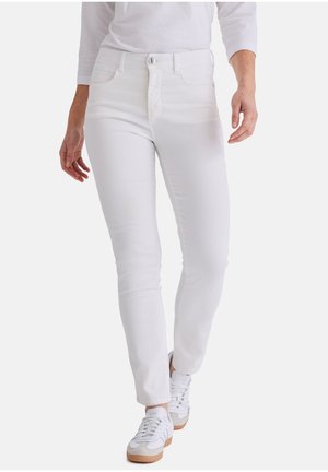 Person trägt weiße Slim-Fit-Jeans und weiße Sneaker mit Gummisohlen und geht vor einem schlichten weißen Hintergrund.