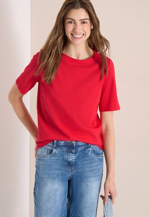 Donna sorridente con capelli castani lunghi che indossa una maglietta rossa a maniche corte e jeans blu, tenendo un capo in denim nella mano destra.