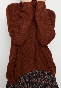 Brown knitted sweater with a loose fit, long sleeves, and a high neck; layered over a floral-patterned skirt. Textured fabric.