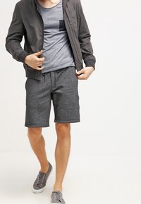 Mężczyzna w ciemnoszarej kurtce z zamkiem, jasnoszarej koszulce, teksturowanych szarych szortach i szarych trampkach idzie do przodu.