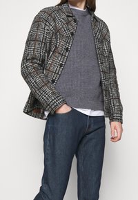 Chaqueta de cuadros abotonada en tonos gris, negro y marrón; tela texturizada, con dos bolsillos en el pecho, combinada con un suéter gris y jeans oscuros.