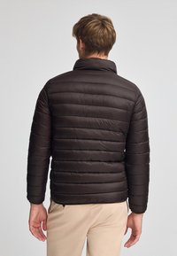 Mörkbrun puffjacka med horisontella kviltade paneler, hög krage och en slät, blank yta, sedd bakifrån.