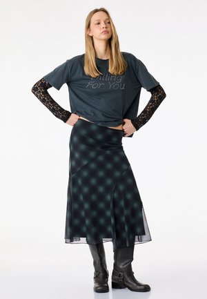 Junge Frau mit schwarzen Spitzen-Ärmeln unter einem dunklen Grafik-T-Shirt, Karo-Midirock und schwarzen Stiefeln bis zur Mitte der Wade steht mit den Händen in der Hüfte.