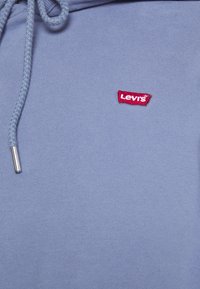 Modrá bavlněná mikina s kapucí, která má texturovaný povrch, stahovací šňůrku na kapuci a výrazné červené logo Levi's na přední straně.