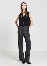 Gilet nero corto con bottoni, abbinato a pantaloni a vita alta grigi a quadri. La modella indossa tacchi neri a punta e posa su uno sfondo neutro.