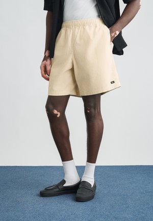 Μπεζ σορτς κοτλέ με ελαστική μέση, πλαϊνές τσέπες και μικρή ετικέτα μάρκας. Συνδυασμένα με λευκές κάλτσες και μαύρα παπούτσια slip-on.