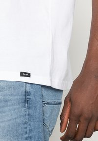 Camisa de algodón blanca con un corte holgado, que presenta una etiqueta negra de "Lee" en el dobladillo, combinada con jeans de mezclilla azul claro. Piel con textura visible.