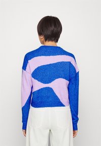 Person med kort mörkt hår iklädd en tröja med blå och lavendel abstrakt mönster och vita byxor, stående framför en enkel ljus bakgrund.