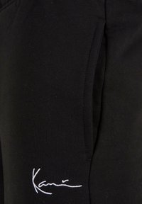 Sorte treningsbukser laget av myk stoff med smal passform, som har en sidelomme og hvit brodert logo foran.