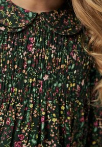 Abito floreale verde scuro con un corpetto plissettato, dotato di un colletto arrotondato e fiori rosa, gialli e bianchi sparsi sul tessuto.