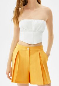 Vit axelbandslös topp med rynkad midja, tillsammans med högmidjade, veckade gula shorts med en framknapp och sidofickor.