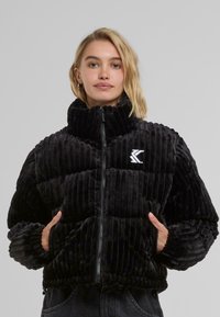Black ribbed puffer jacket made of soft velvet. Features a high collar, zip front, and elastic cuffs. White logo accent on the chest.