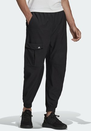 Mann trägt schwarze Jogginghosen mit seitlicher Cargotasche und schwarzen Turnschuhen, steht mit einer Hand in der Tasche.