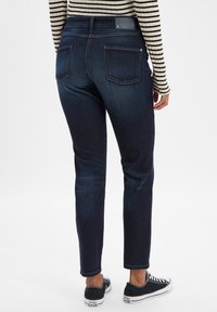 Jeans de mezclilla oscura con un ajuste slim, dos bolsillos traseros y una cinturilla texturizada. Combinados con zapatillas de lona en blanco y negro.
