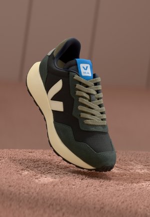 Ciemnozielony i czarny sneaker Veja z beżową podeszwą i sznurówkami, z białym logo "V" oraz niebieską metką na języku, na teksturowanej powierzchni.