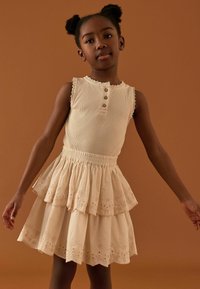 Débardeur beige à côtes avec trois boutons, associé à une jupe à volants superposés ornée de détails en œillets. Texture lisse, design féminin.
