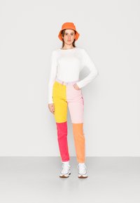Camisa de manga longa de algodão, branca sólida. Calças de veludo cotelê em várias cores com painéis rosa, amarelo e laranja. Chapéu de balde laranja. Ténis brancos.