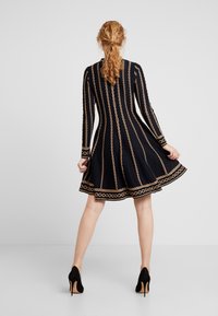 Robe noire à manches longues, ornée de rayures à motifs de chaînes dorées. Jupe évasée et détails décoratifs aux poignets. Portée avec des talons noirs.