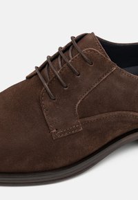 Brauner Herrenschuh aus Wildleder mit geschlossener Schnürung, detaillierter Nahtführung und dunkler Gummisohle vor weißem Hintergrund.