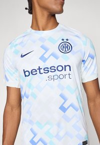 Un maillot de football blanc à manches courtes avec des motifs géométriques bleu clair, arborant le logo "betsson.sport" et un badge d'équipe brodé avec deux étoiles.