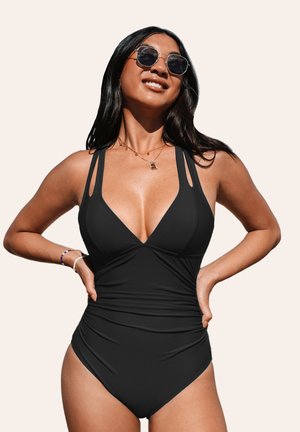 Maillot de bain noir d'une pièce avec un décolleté plongeant, des bretelles larges et un tissu froncé sur le côté. La mannequin porte des lunettes de soleil rondes et des bijoux simples.