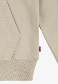 Felpa beige con tessuto testurizzato, dotata di tasca a marsupio, polsini a costine e un piccolo'etichetta rossa sul lato.